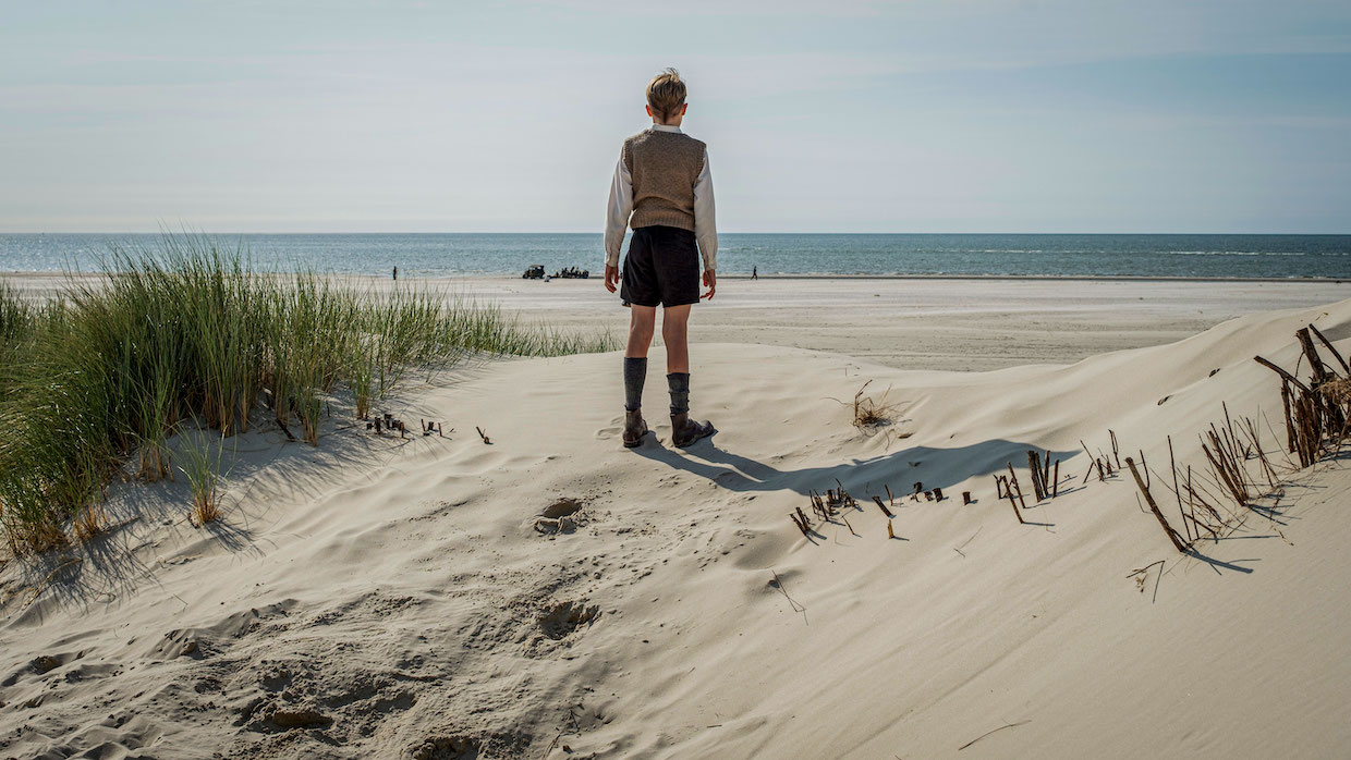 A boy stands on a sandy shore in the summer. He wears 1940s-era clothes, including shorts, a button-up, sweater vest and knee-high socks. His back faces us as he looks out at the blue water on a sunny day.