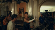 A woman in a navy ballgown and white opera gloves directs a film set. She speaks to her crew in between takes, where she is also starring as the lead role.