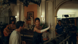 A woman in a navy ballgown and white opera gloves directs a film set. She speaks to her crew in between takes, where she is also starring as the lead role.