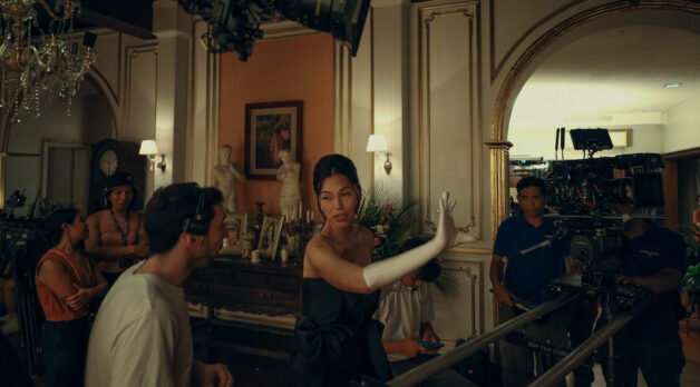 A woman in a navy ballgown and white opera gloves directs a film set. She speaks to her crew in between takes, where she is also starring as the lead role.