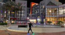 A man wears all black clothing and walks around downtown Los Angeles. The soles of his shoes glow blue and green.