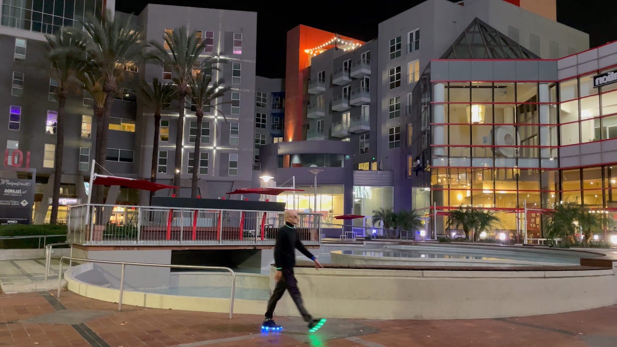 A man wears all black clothing and walks around downtown Los Angeles. The soles of his shoes glow blue and green.