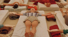 Rows of people lay blindfolded on mattresses. Their arms are stretched out to the side and they lay on white sheets. At the center, a woman with honey-colored hair wears a top with gray and white gradient vertical stripes. She wears cottons shorts with tan and white stripes.