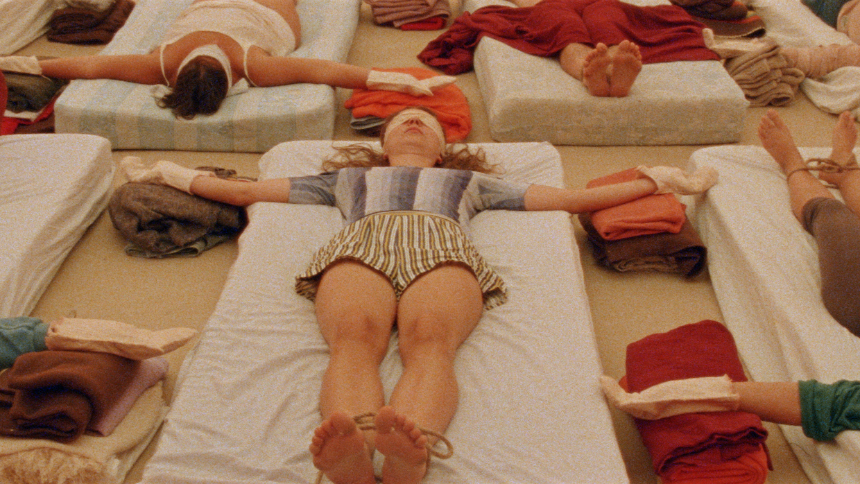 Rows of people lay blindfolded on mattresses. Their arms are stretched out to the side and they lay on white sheets. At the center, a woman with honey-colored hair wears a top with gray and white gradient vertical stripes. She wears cottons shorts with tan and white stripes.