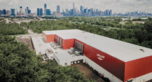 A long, rectangular, red building containing a soundstage is captured via drone, with the NYC skyline lining the background.