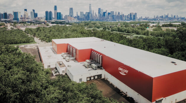 A long, rectangular, red building containing a soundstage is captured via drone, with the NYC skyline lining the background.