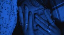 A close-up of a young woman with an eyebrow piercing and brown bangs covering her face with her hands. She is washed in blue light.