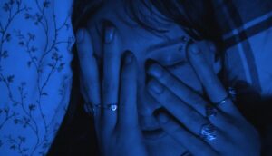A close-up of a young woman with an eyebrow piercing and brown bangs covering her face with her hands. She is washed in blue light.