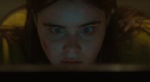 A close-up of a young woman with brown hair. She stares wide-eyed as she looks at a computer screen.