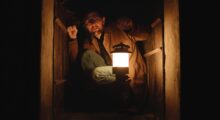 A man wearing a tan jacket, jeans, and glasses crouches in a dumbwaiter. He holds a lamp out in front of him, otherwise engulfed in darkness.