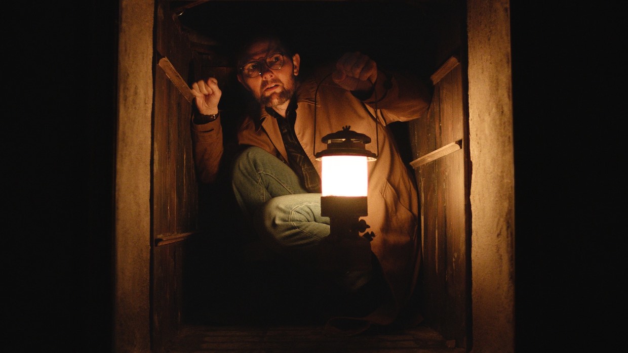A man wearing a tan jacket, jeans, and glasses crouches in a dumbwaiter. He holds a lamp out in front of him, otherwise engulfed in darkness.