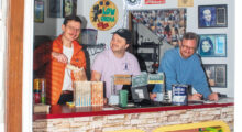 Three men stand behind a concession stand with a collage of cinematic ephemera hung on the walls behind them. One on the left wears an orange parka and scoops popcorn into a bag. The one in the center wears an off-pink shirt and smiles, looking at the man scooping popcorn. A man in a blue pullover leans on the counter and smiles.