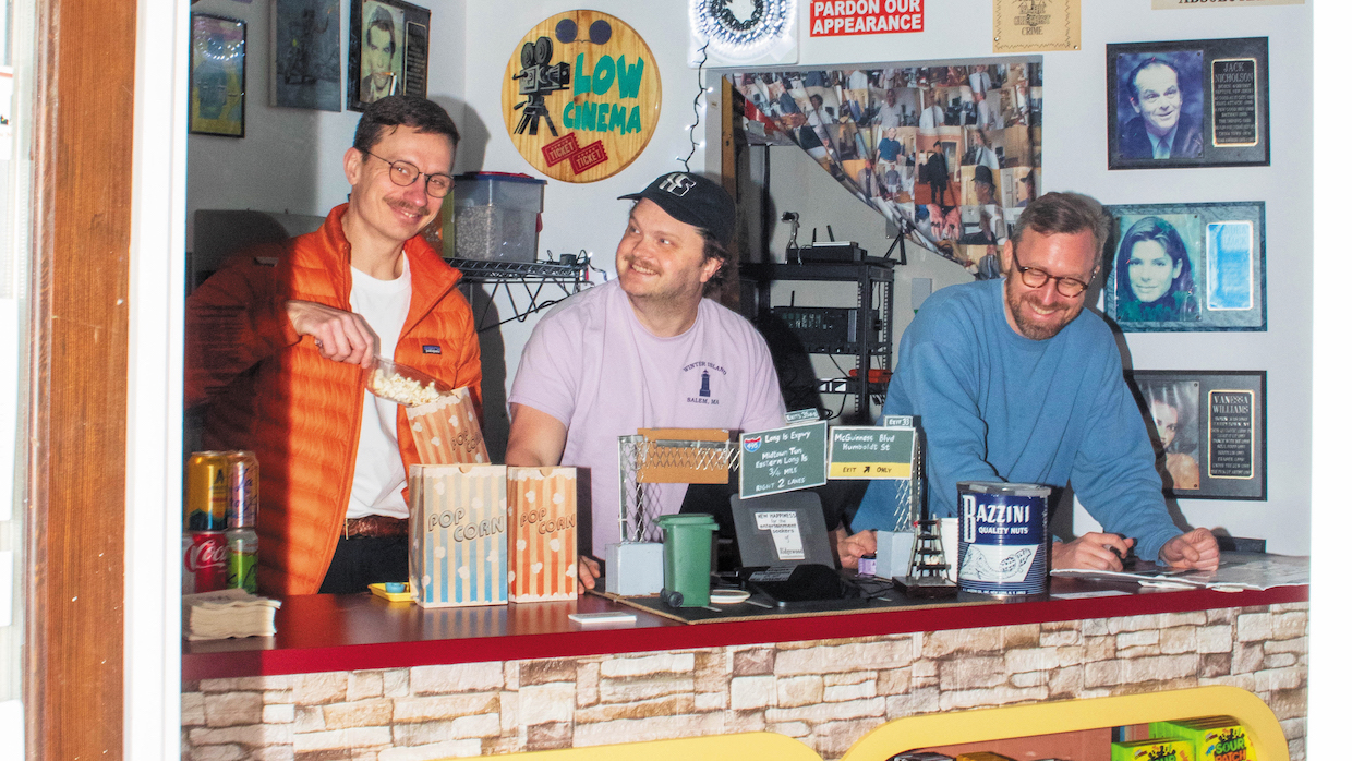 Three men stand behind a concession stand with a collage of cinematic ephemera hung on the walls behind them. One on the left wears an orange parka and scoops popcorn into a bag. The one in the center wears an off-pink shirt and smiles, looking at the man scooping popcorn. A man in a blue pullover leans on the counter and smiles.