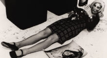 A black and white photo of a woman wearing a plaid dress, black shoes and heavy make-up laying on a concrete floor. A photo resembling her lays on the ground to her left.