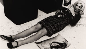 A black and white photo of a woman wearing a plaid dress, black shoes and heavy make-up laying on a concrete floor. A photo resembling her lays on the ground to her left.