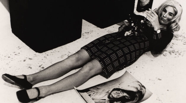 A black and white photo of a woman wearing a plaid dress, black shoes and heavy make-up laying on a concrete floor. A photo resembling her lays on the ground to her left.