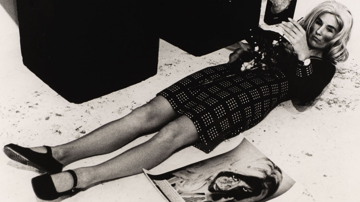 A black and white photo of a woman wearing a plaid dress, black shoes and heavy make-up laying on a concrete floor. A photo resembling her lays on the ground to her left.