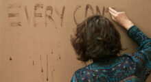 A woman has her back facing the camera. She uses a liquid to write "every contact" on a blank tan canvas.