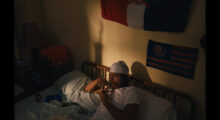 A young man smokes weed from a bowl in his bed, the setting sun casts shadows on the wall, where a Dominican flag hangs above his bed.