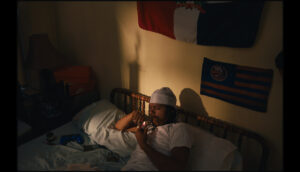 A young man smokes weed from a bowl in his bed, the setting sun casts shadows on the wall, where a Dominican flag hangs above his bed.