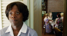 A Black woman in a maid's uniform stands to the side of an entryway looking glum and defeated. Through the entryway, a while family eats dinner.