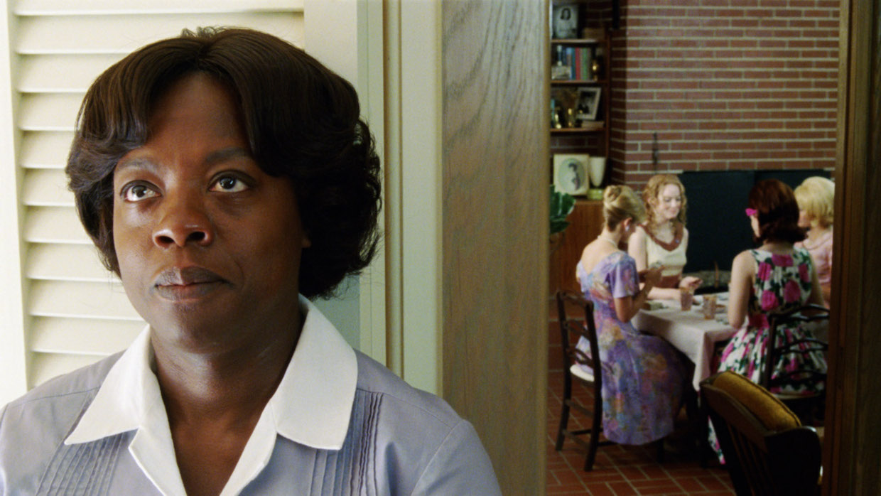 A Black woman in a maid's uniform stands to the side of an entryway looking glum and defeated. Through the entryway, a while family eats dinner.