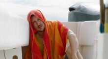 A man sits on a white boat with a orange and pink towel draped over his head.