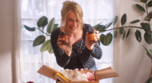 A man in drag wears a blond wig in an updo. She is wearing a long sleeve black shirt and stands in front of a house plant. She hovers above an open box and holds two jars of jam in her hands.