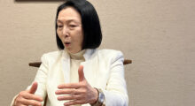 An older Japanese woman with a black bob and wearing a white top and blazer gesticulates as she speaks at a wooden table.