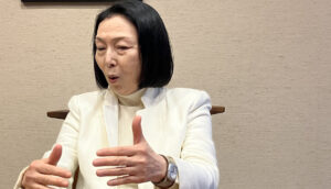 An older Japanese woman with a black bob and wearing a white top and blazer gesticulates as she speaks at a wooden table.