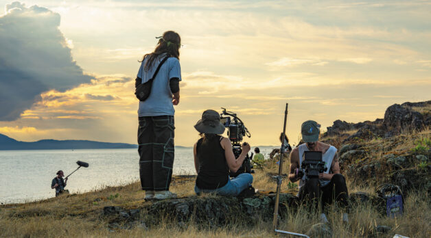 A film crew sets up on the edge of a tall hill on Vancouver Island. The sun sets in the sky behind them.