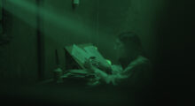 A woman sits in a sparse medieval room and writes on parchment. She is cast in green light as a beam shines on her from a window.