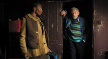 A Black woman wears a brown vest over a yellow button-up and faces an older white man, who wears a denim jacket and a blue, green and black striped sweater and leans against a doorway in a room made of chipped, dark wood.