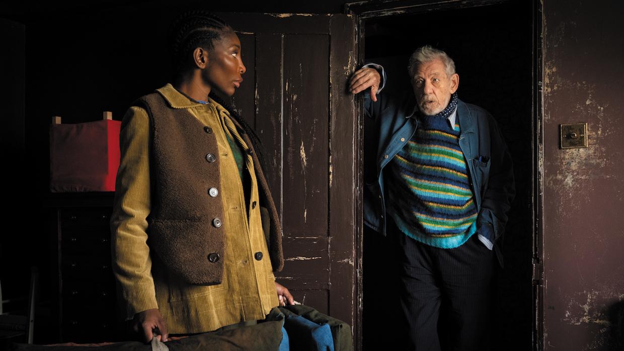 A Black woman wears a brown vest over a yellow button-up and faces an older white man, who wears a denim jacket and a blue, green and black striped sweater and leans against a doorway in a room made of chipped, dark wood.