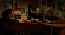 Two older Italian men and a younger one sit in a dimly lit bar, all clutching drinks and engaged in conversation.