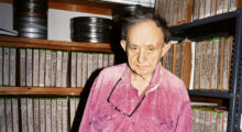 An elderly man wears a bright pink shirt, glasses hanging around his neck. He stands among shelves of film prints in an archive.