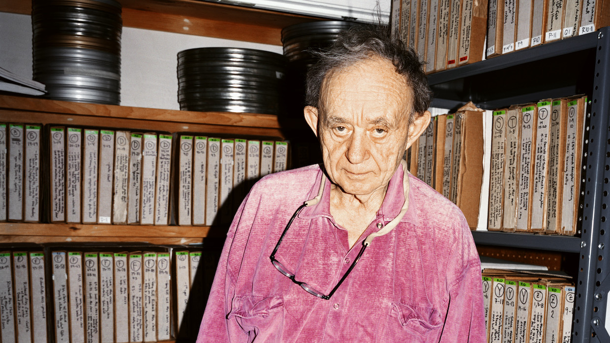 An elderly man wears a bright pink shirt, glasses hanging around his neck. He stands among shelves of film prints in an archive.