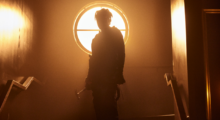 A masked figure is backlit by a sun-filled window, casting him in a shadow.