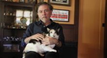 A white older man sits in a chair in an office with comic art on the walls. He wears a blue button-up with the sleeves rolled up. Nestled in his lap is a handsome calico cat.