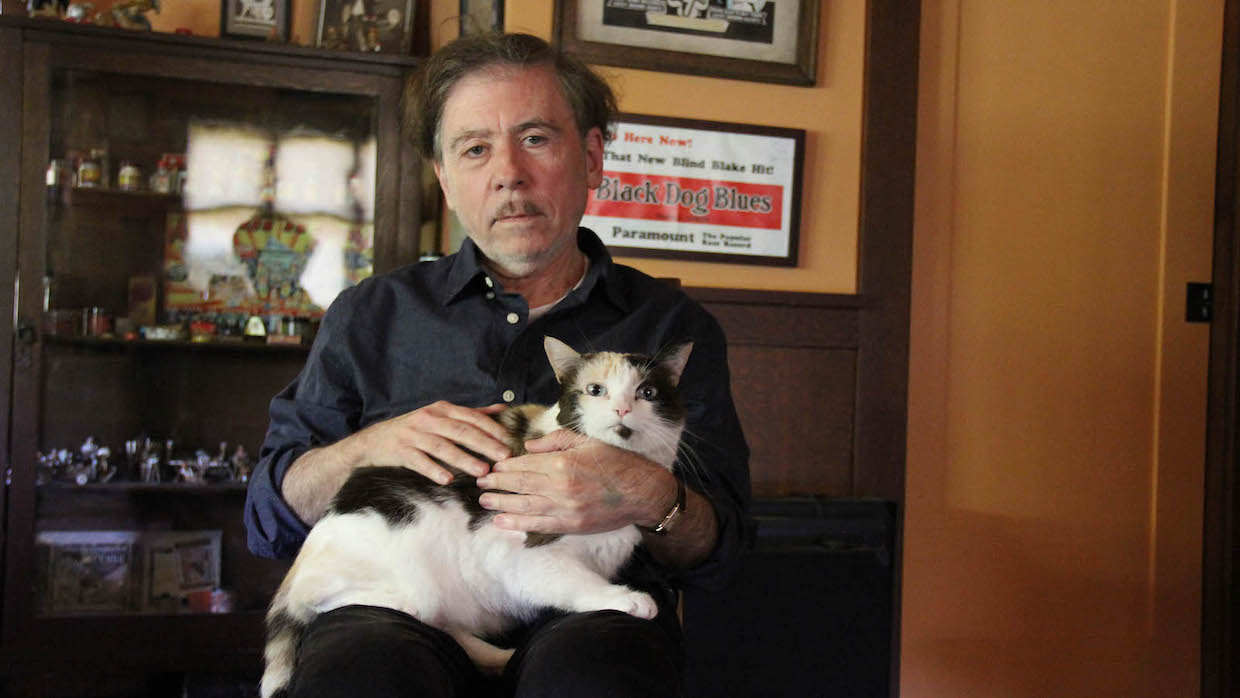 A white older man sits in a chair in an office with comic art on the walls. He wears a blue button-up with the sleeves rolled up. Nestled in his lap is a handsome calico cat.