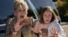 An older woman with a blonde beehive scrams and puts her hands out in front of her. A younger woman with a strawberry blonde bob looks confused and also holds her hands in front of her.
