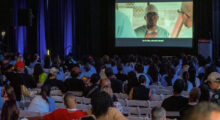 An audience of women inmates in blue uniforms and civilians watch a film on a screen.