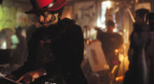 A Black man with mutton chops and a tall red hat points to a camera he is holding down by his waist. A filmmaking crew is out of focus in the background behind him.