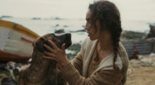 A woman with long, brown hair in a braid wears a tan cardigan. She kneels on a beach and her hands hold a brown dog's face in her hands.