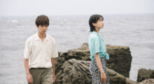 A young Japanese man and woman stand by a jetty near the sea. He wears a white shirt and tan pants, while she dons a mint blouse and black long skirt with small white flowers printed on it.
