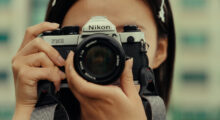 A woman takes a photo with a vintage Nikon camera, obscuring her face.