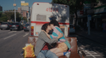 A young couple kiss on a tan leather couch that is in the middle of the street. A bus is on the road in front of them.
