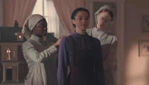 A young woman wears a modest purple dress. Two other young women, wearing taupe modest dresses with matching head coverings, stand behind her and help zip up the garment.