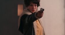 A woman with short, curly hair holds a gun out in front of her.