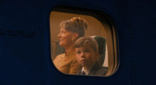 A young boy wearing a suit and tie looks out an airplane window. His mother, wearing a taupe tweed coat, looks straight ahead.